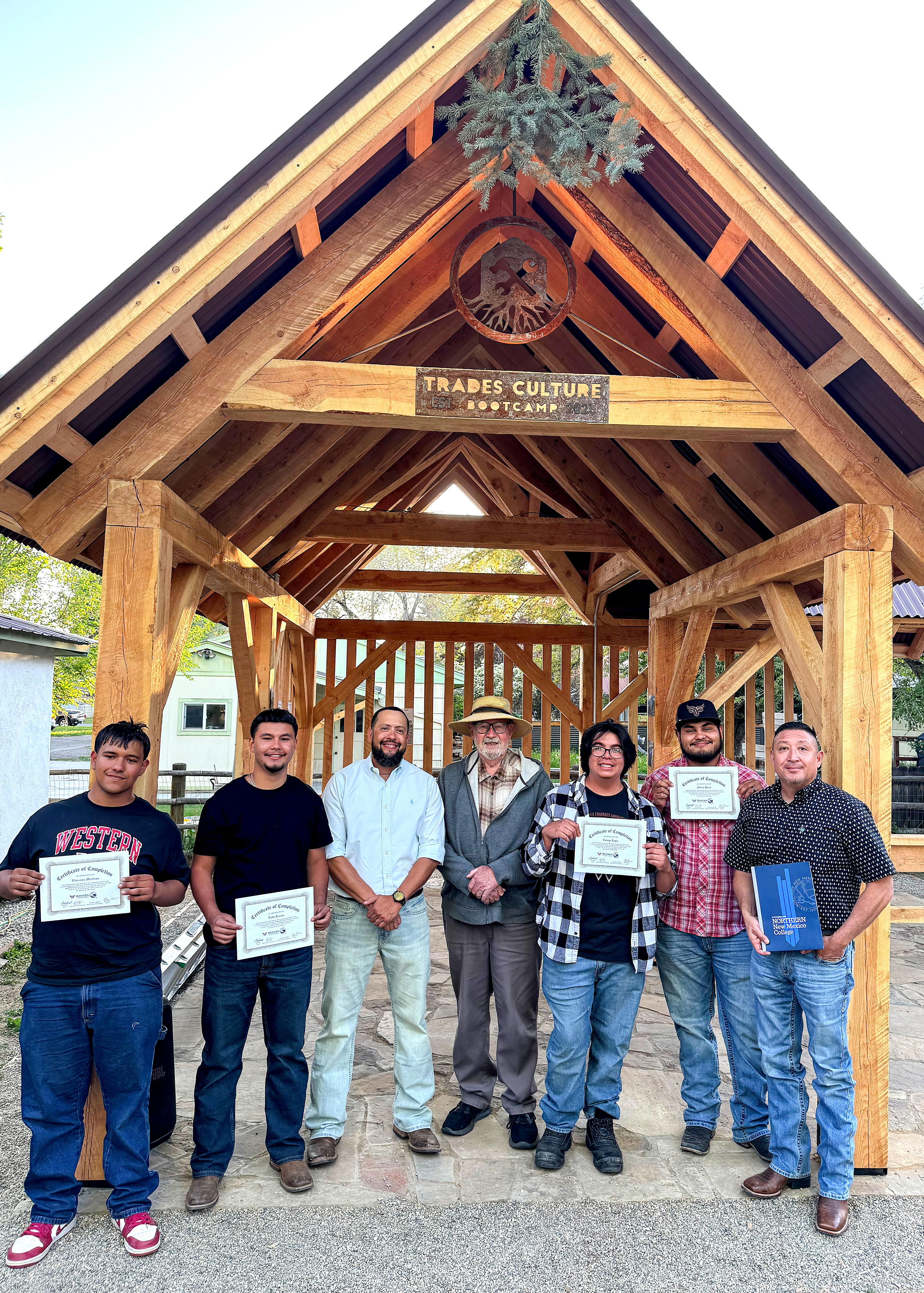 A photo of the timber framed gazebo with the NNMC Bootcamp crew in front of it. From left: Edwardo Martinez, Luka Torrez, Marvin Romero, Butch Clark, George Lopez, Mario Perez and Joe Padilla. A spruce branch hangs from the peak of the structure with the plaques created by the students below it.